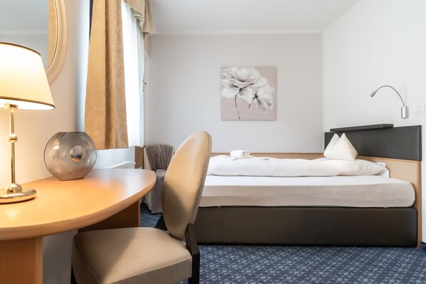 A hotel room with a bed, a desk with a lamp, a chair, and a wall-mounted artwork.