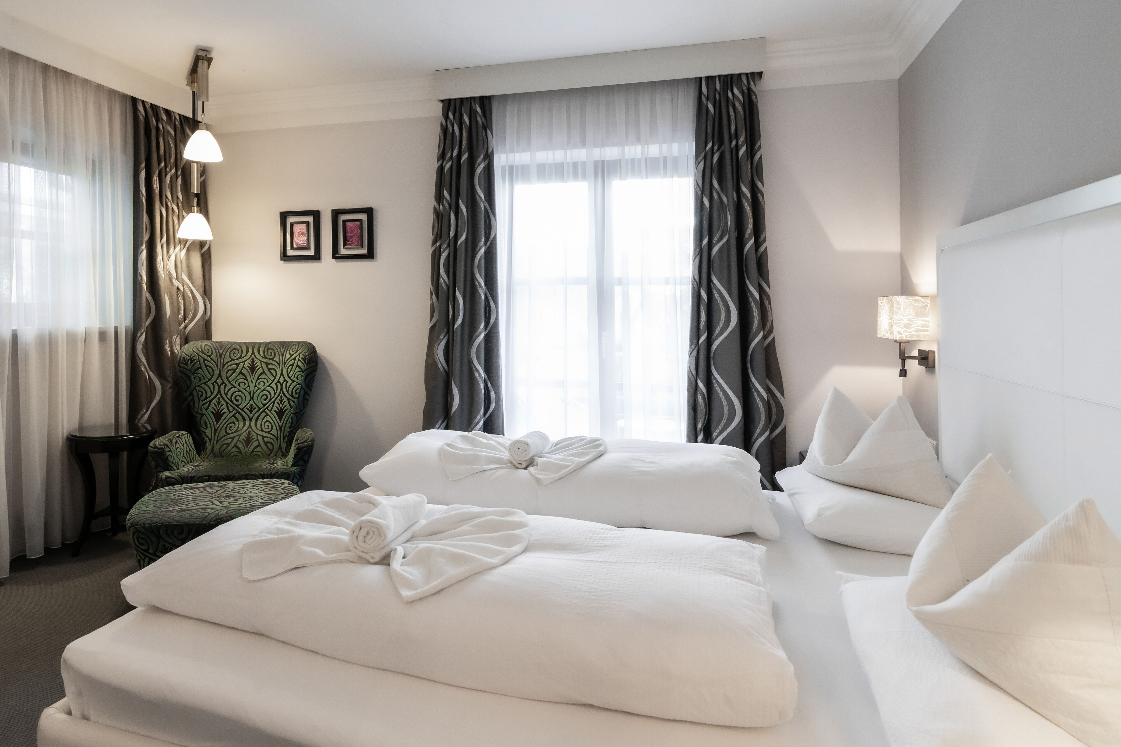 A hotel room with two beds, a green patterned armchair, a side table, and a lamp.