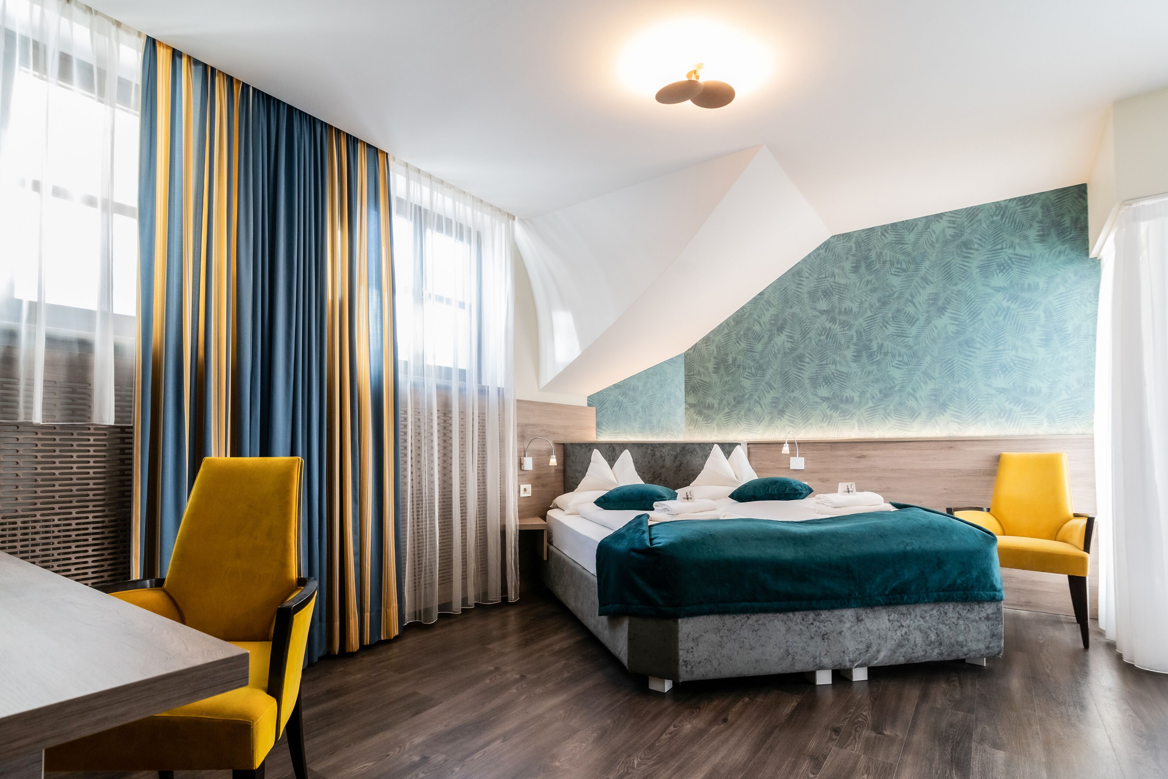 Una habitación de hotel moderna con una cama, dos sillones amarillos, una mesa y un ventanal con cortinas.