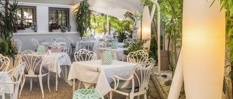An outdoor patio with white tables and chairs, surrounded by greenery and plants.
