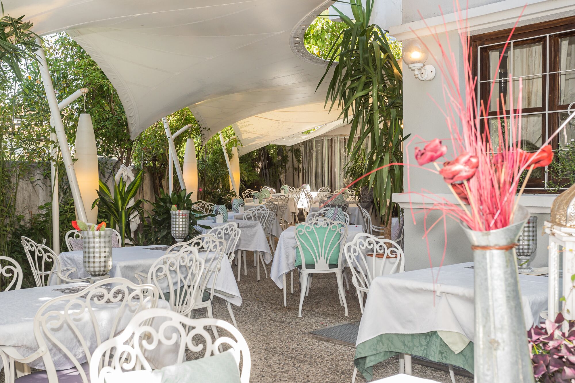 Un espace de restauration en plein air avec des chaises et des tables en métal blanc, protégé par un auvent blanc et entouré de verdure.
