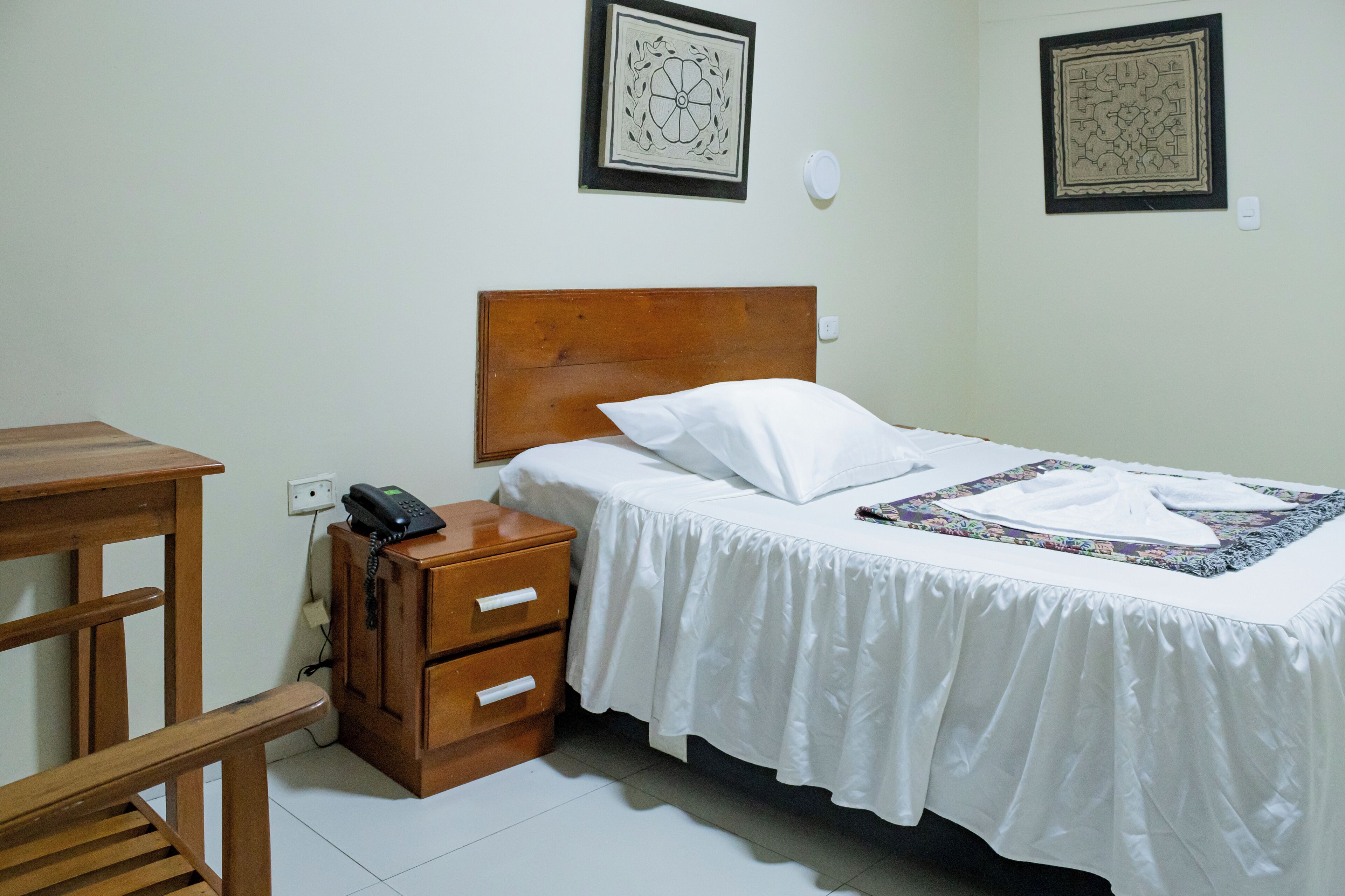Un dormitorio ordenado con una cama con cabecera de madera, una mesita de noche con un teléfono y dos interruptores de luz en la pared.