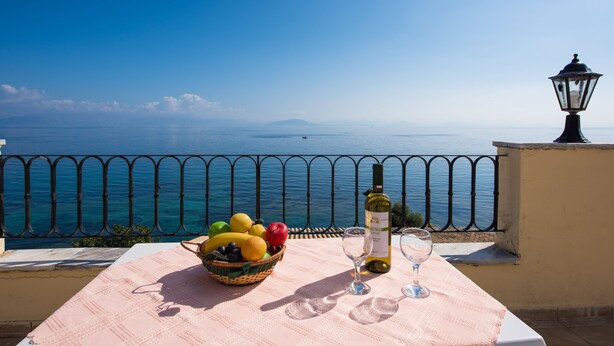 Une table avec un panier de fruits, une bouteille de vin et des verres, sur un balcon donnant sur la mer.