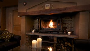 A cozy living room with a fireplace, two candles on a glass table, and a stack of firewood.