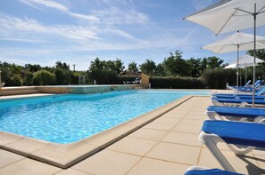 Una piscina con agua cristalina azul, rodeada de sillas de descanso y sombrillas, con un edificio pequeño al fondo.