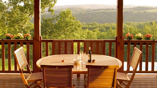 A wooden deck with a round table and chairs, overlooking a forested valley.