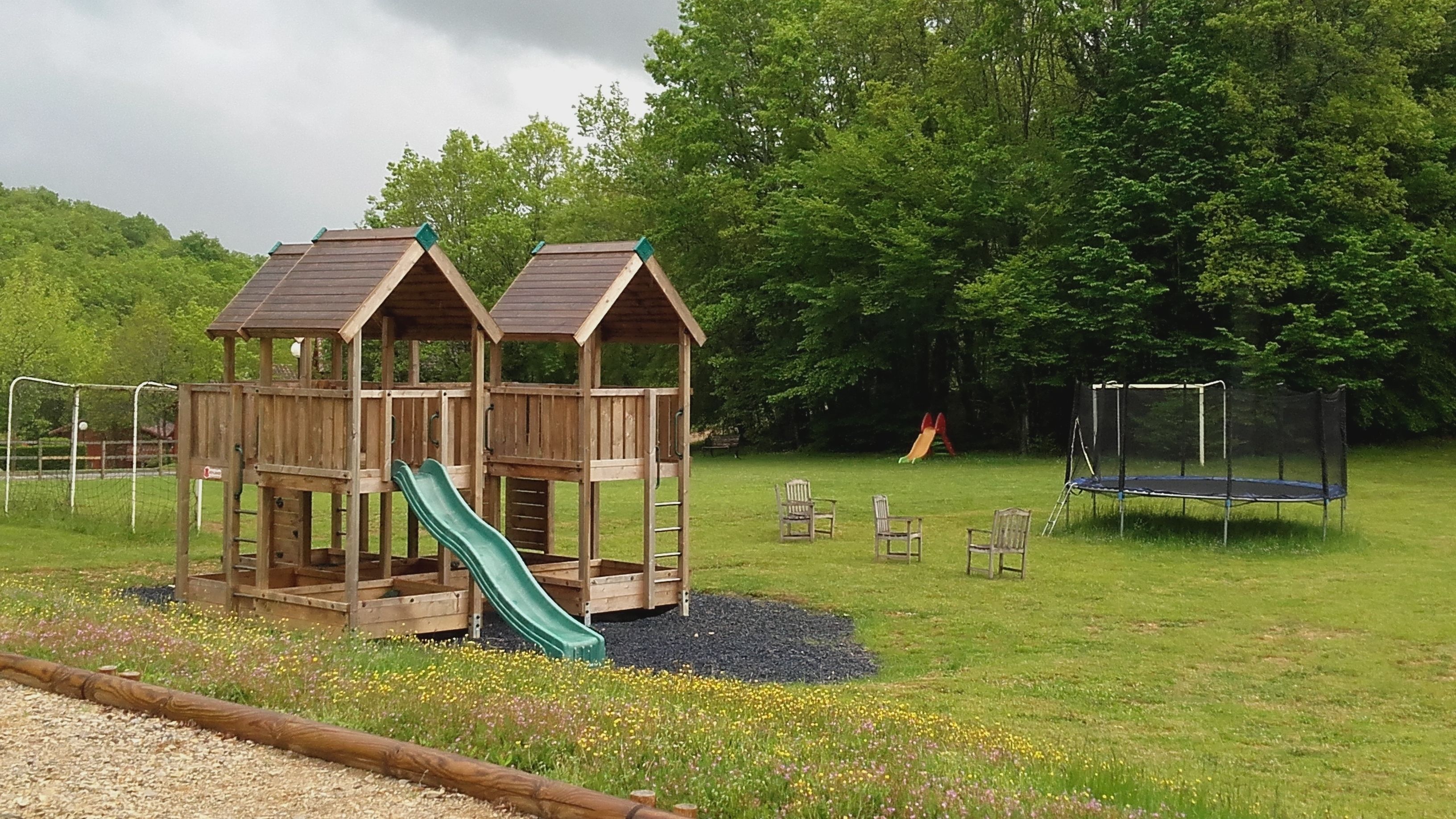 A wooden play structure with a slide and a climbing ladder, surrounded by a grassy field and trees.