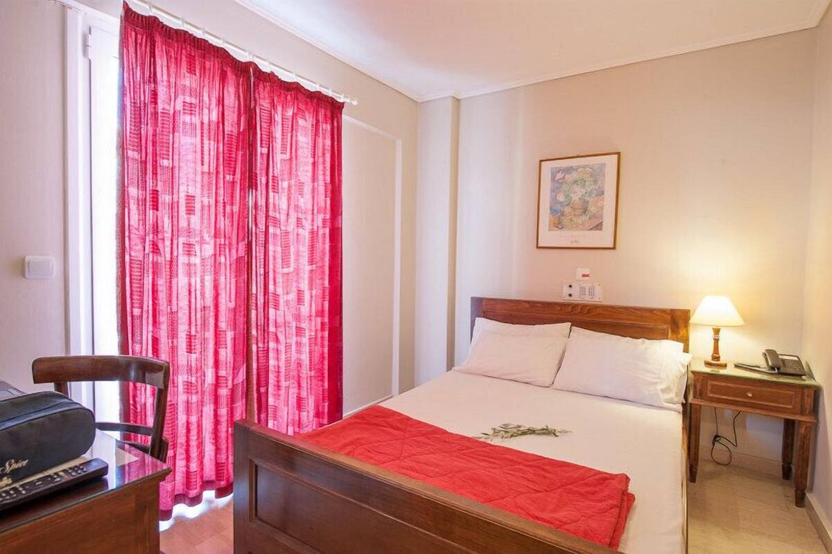 A hotel room with a bed, a bedside table with a lamp, a chair, and a window with red curtains.