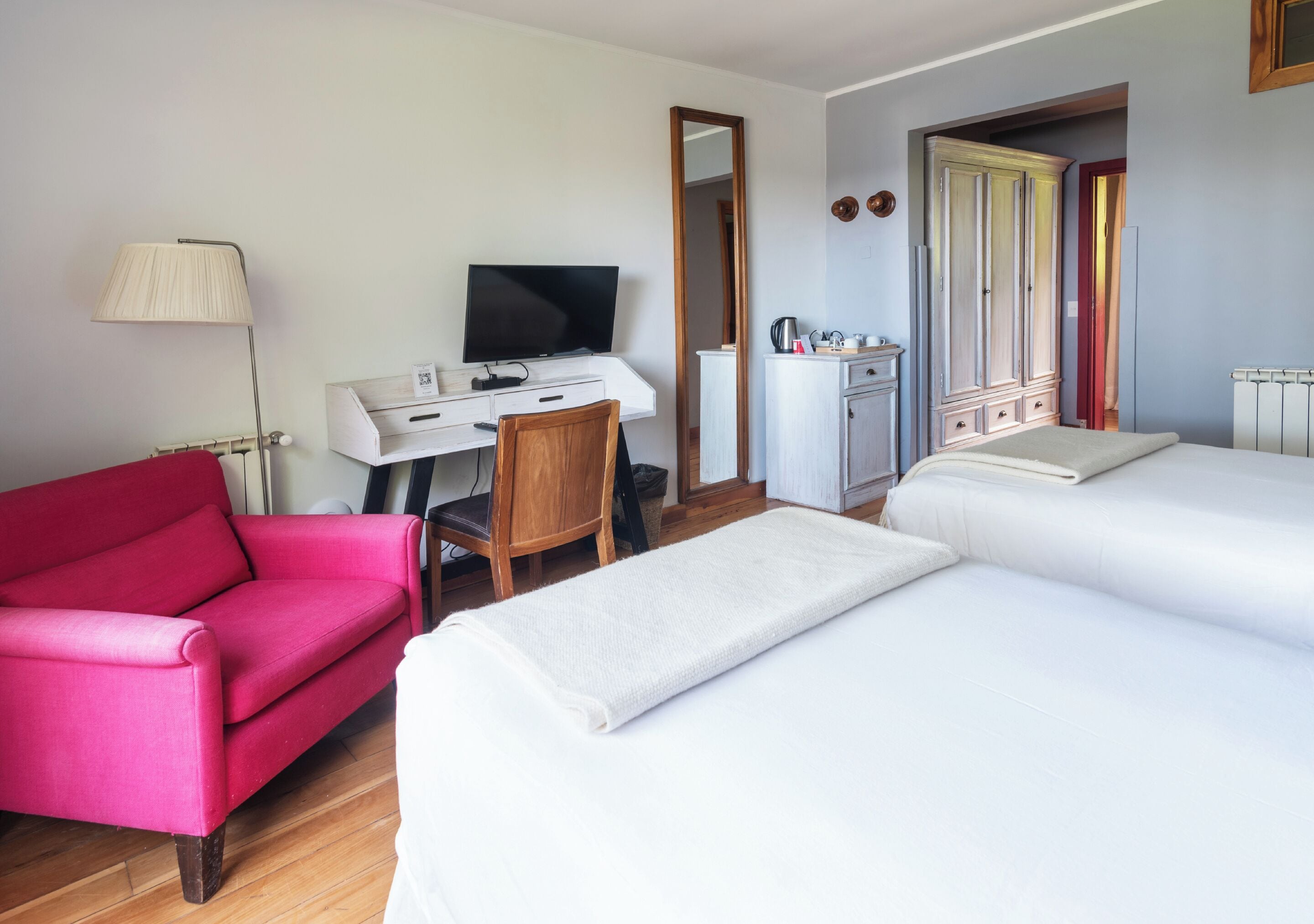 Une chambre d’hôtel équipée d’un bureau, d’une télévision, d’un fauteuil rouge, de deux lits et d’une armoire.