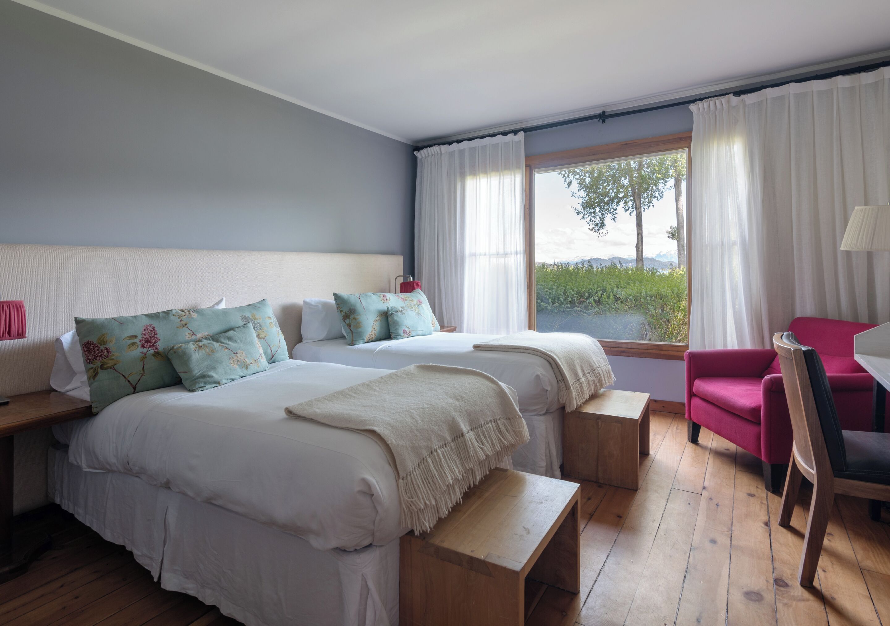 Quarto com duas camas, um banco de madeira, uma cadeira vermelha e vista para o verde pela janela.
