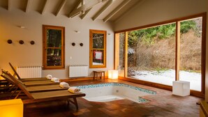 A spa room with a wooden bench, a small table, a bathtub, and large windows offering a view of the outdoors.