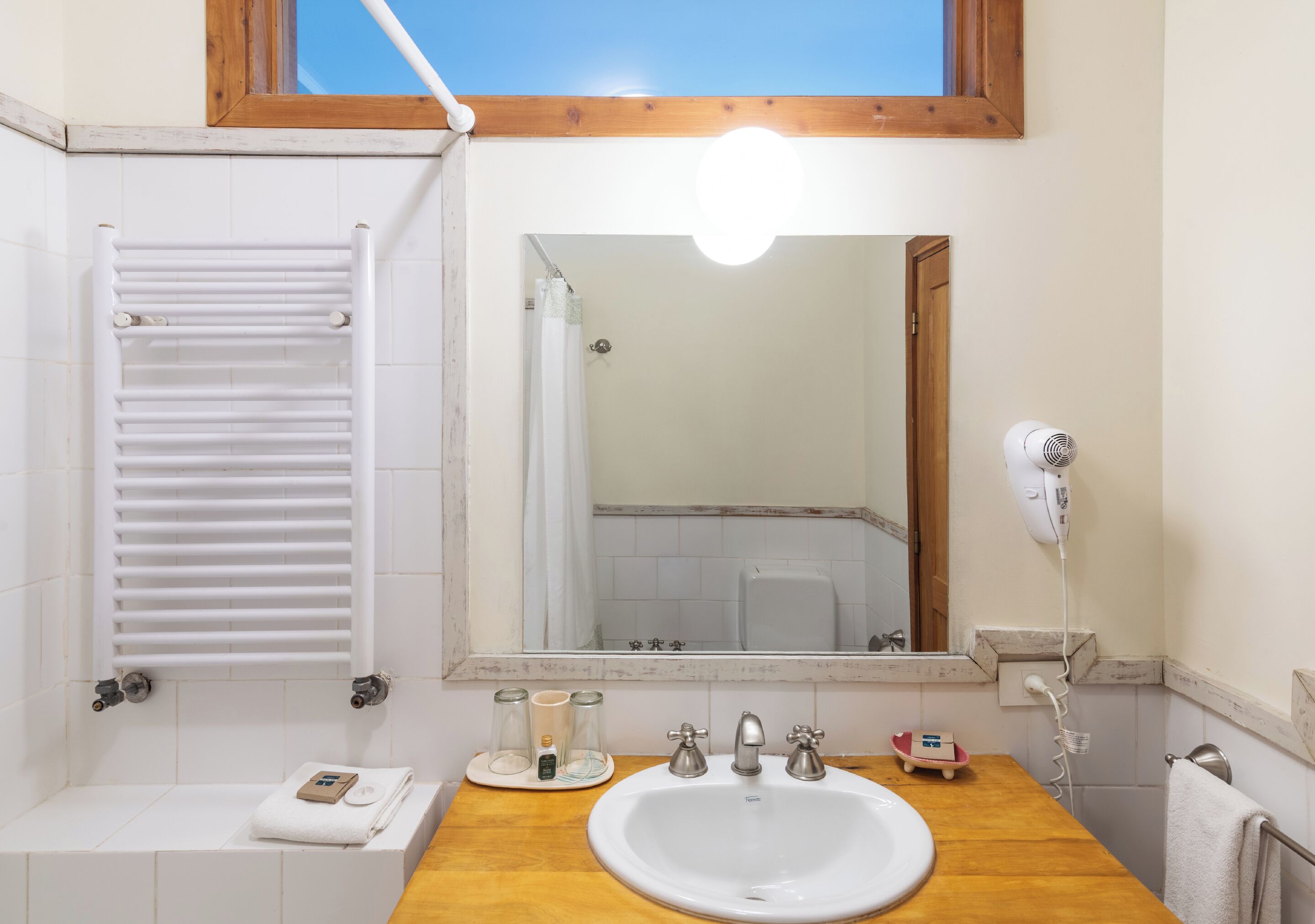 Un bagno con un lavandino bianco, due rubinetti, uno specchio, un portasciuga-asciugamani, una tenda da doccia e un asciugacapelli.