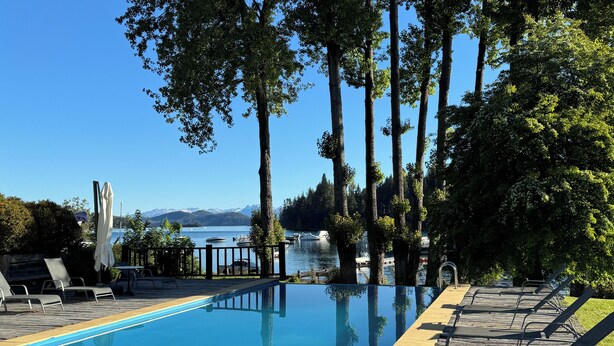 A clear blue pool with lounge chairs and a view of the lake and mountains.