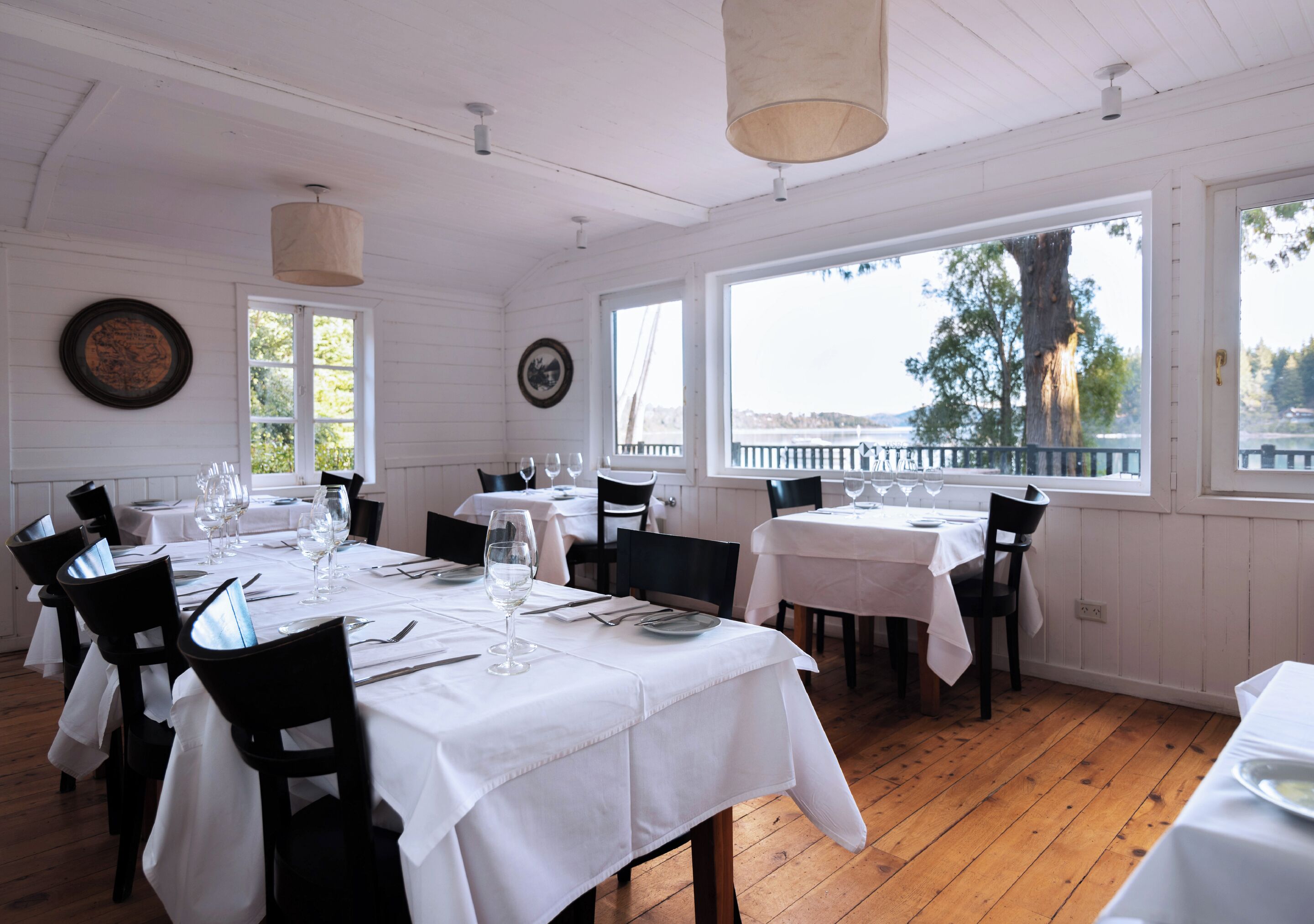 A dining area with white tablecloths, black chairs, and large windows offering a view of the outdoors.