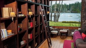 A cozy room with a wooden bookshelf, a ladder, a sofa, and a view of a lake with boats.