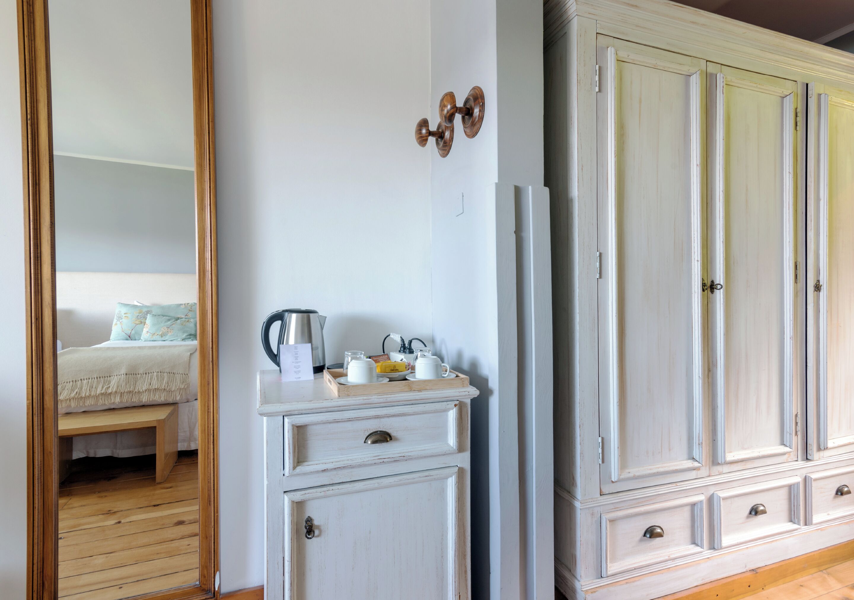 Un dormitorio con suelo de madera, una cama con edredón blanco, una mesita de noche con tetera, tazas y una bandeja, y un armario con un espejo encima.