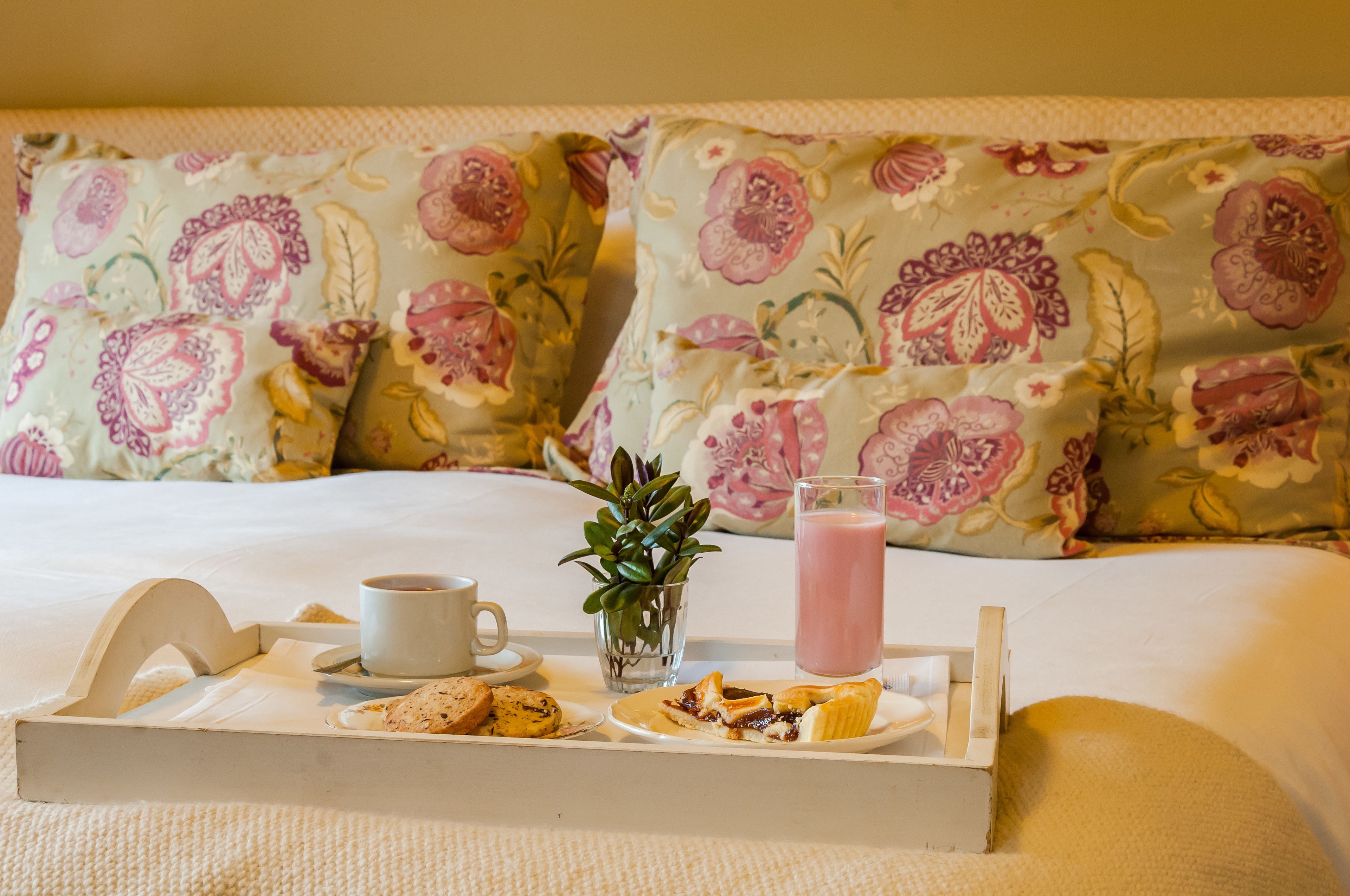 A neatly made breakfast tray with pastries, a cup, a glass of juice, and a small plant on a bed with floral pillows.