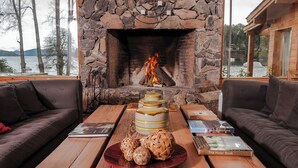 A stone fireplace with a fire, a wooden table with books and woven balls, and a view of a lake and mountains.