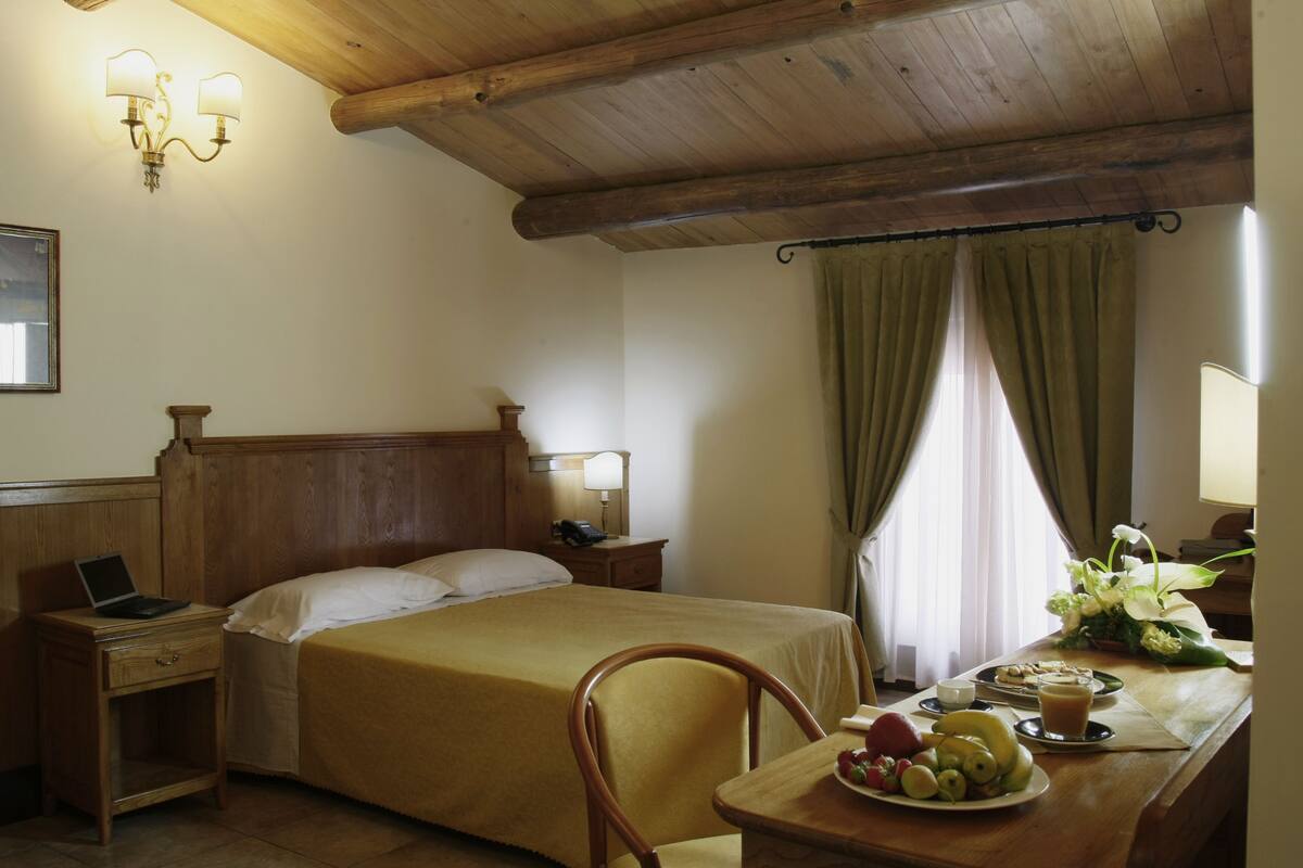 Hotelkamer met een houten bed, nachtkastje, lamp en een ontbijttafel met fruit en een koffiekop.