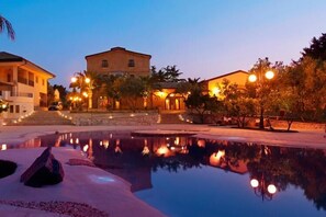 A tranquil pool area with a reflection of buildings and street lamps, surrounded by palm trees and a clear sky.