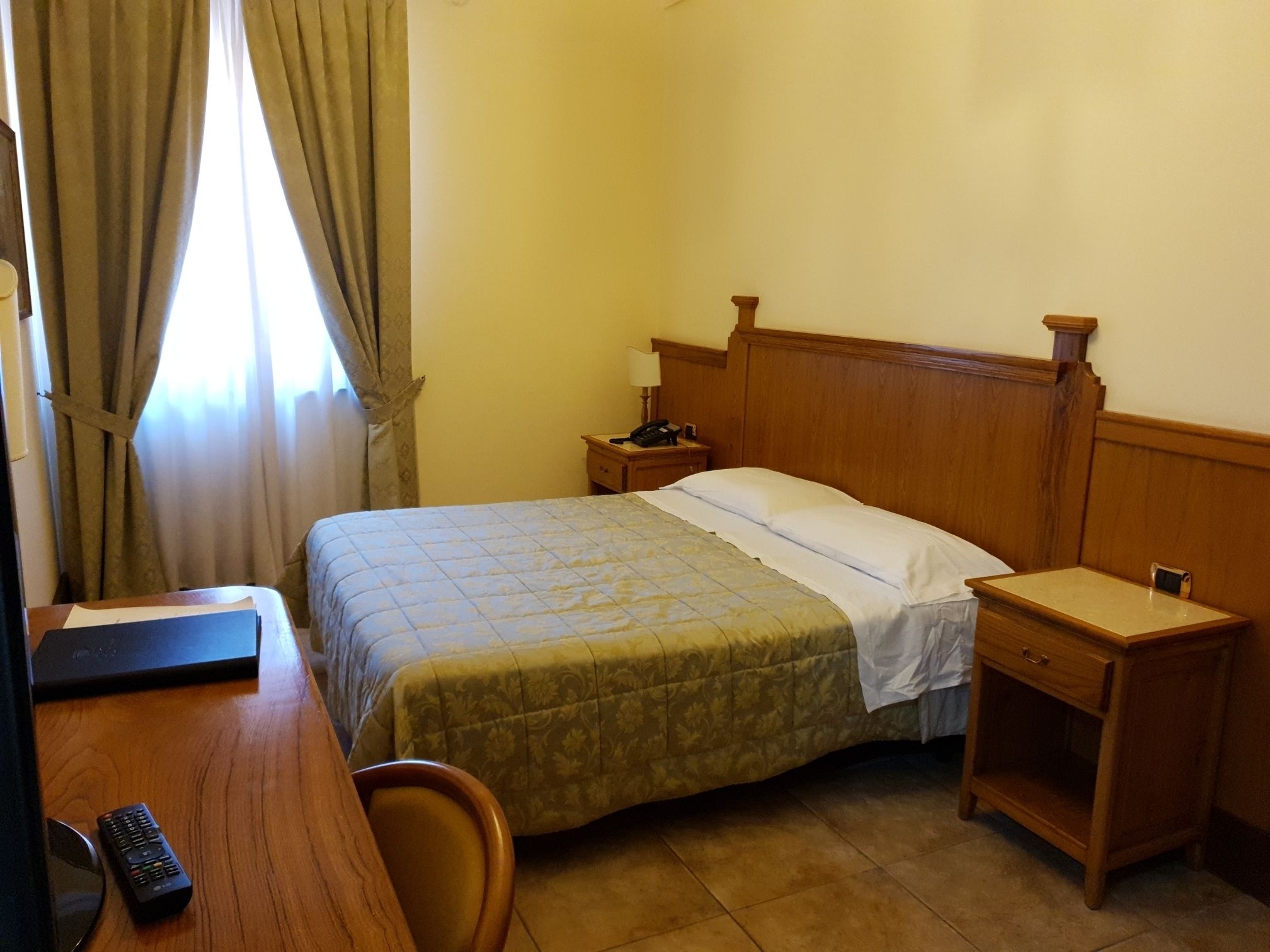 Habitación de hotel con cama, mesita de noche de madera, silla y escritorio con control remoto.