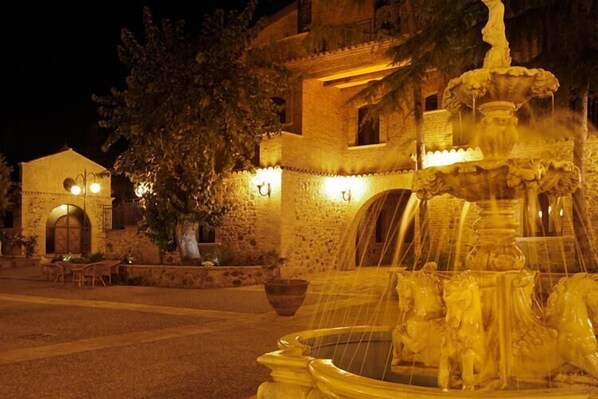 A stone building with a fountain featuring horse sculptures, surrounded by outdoor seating and illuminated by warm lighting.