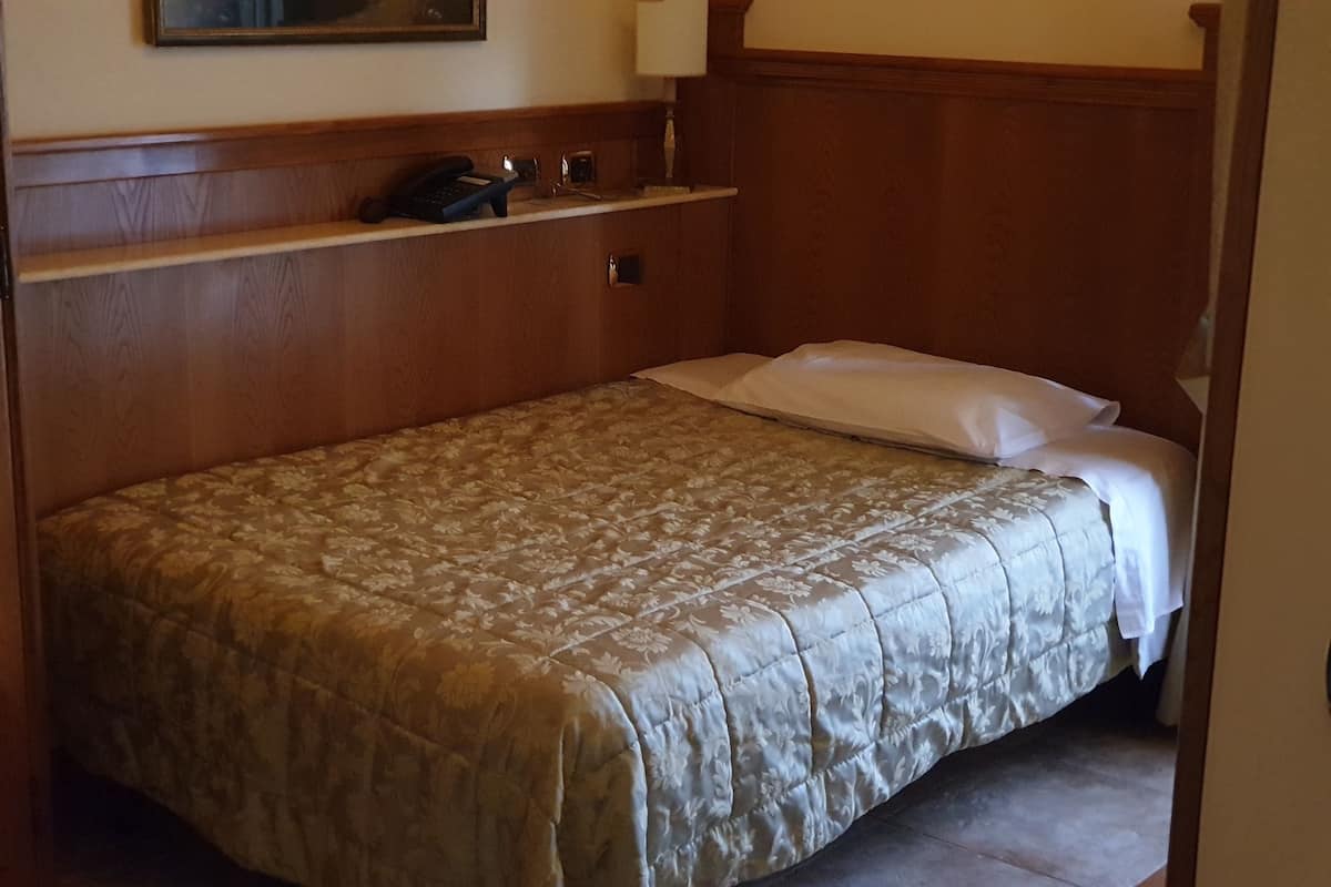 Een netjes opgemaakt bed met een bloemenprint deken en kussens, een houten hoofdbord, en een telefoon op een plank erboven.