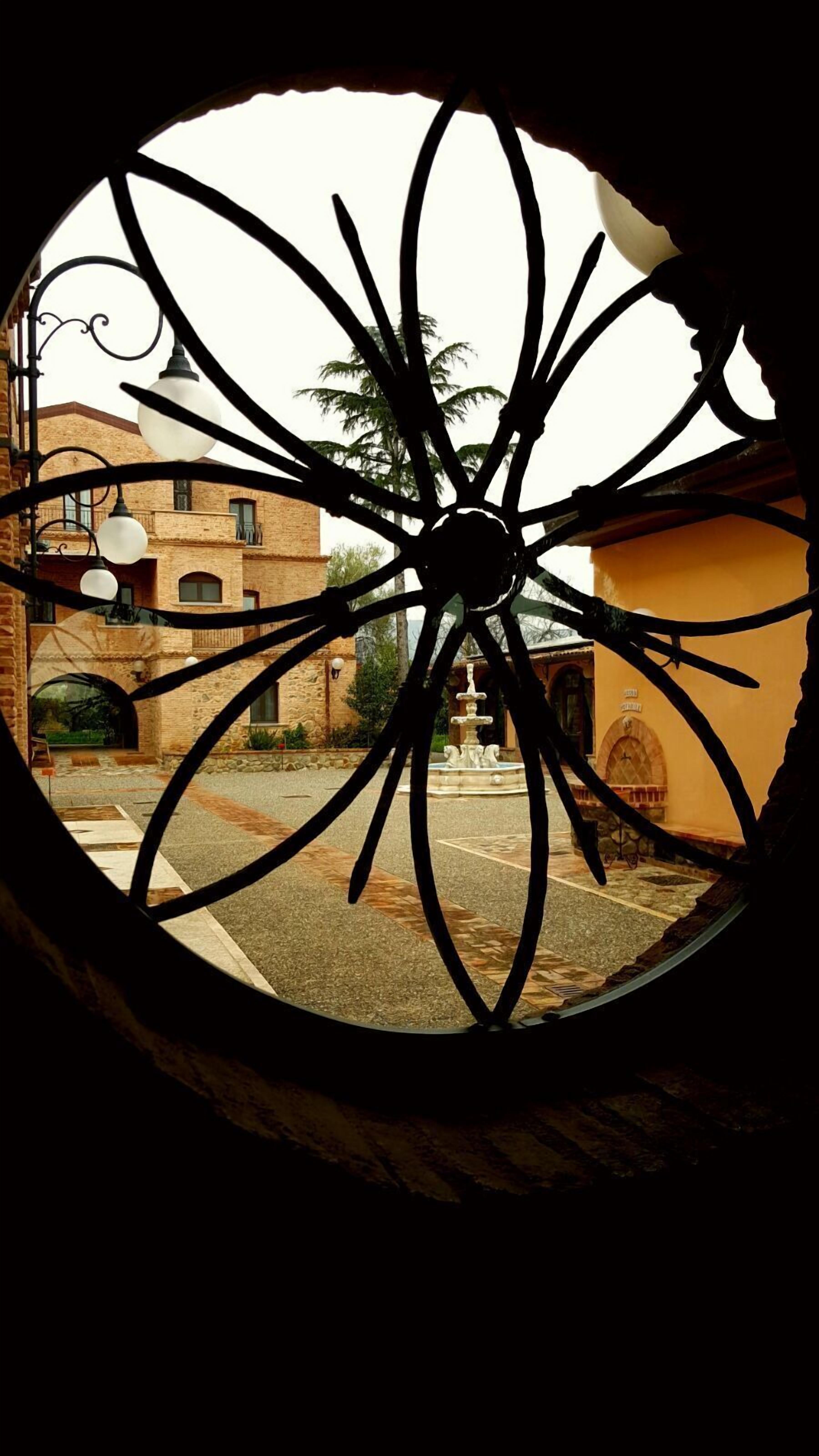 A view through a circular window with a decorative metal frame, featuring a street scene with buildings, a fountain, and a palm tree.