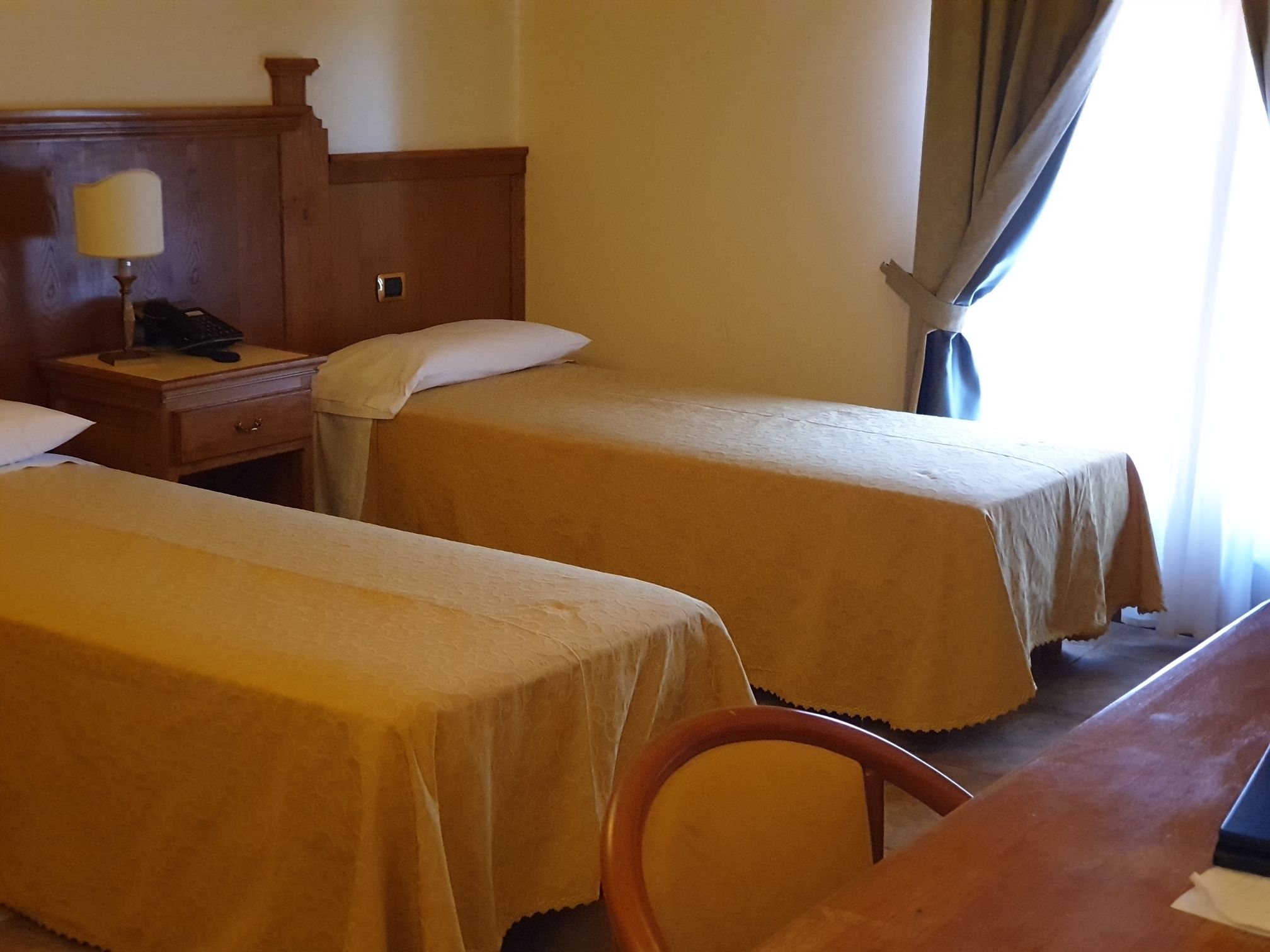 Habitación de hotel con dos camas individuales, una mesita de noche de madera con lámpara, un escritorio y una silla.