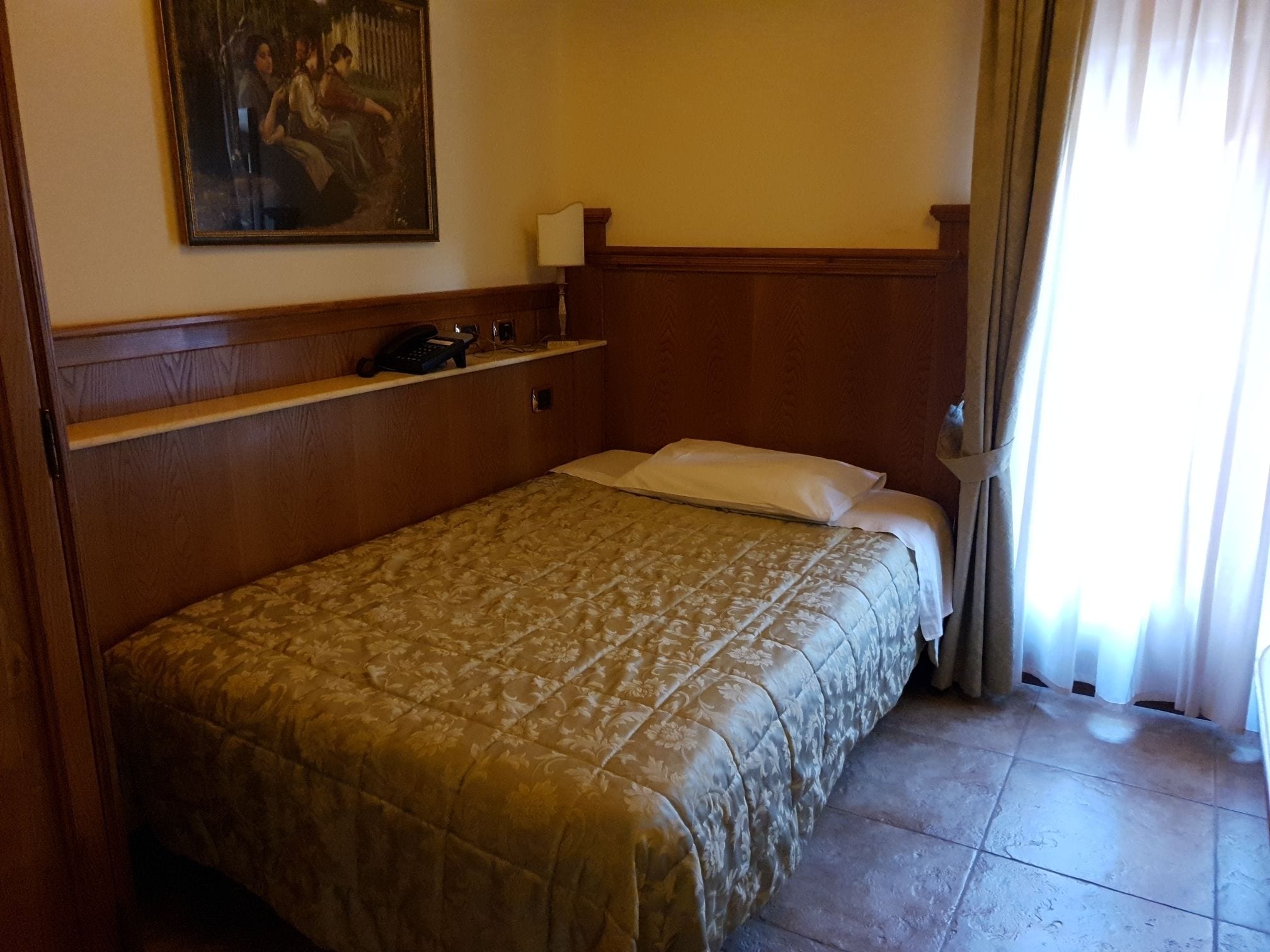 Una habitación de hotel con cama, teléfono, un cuadro en la pared y ventana con cortinas.