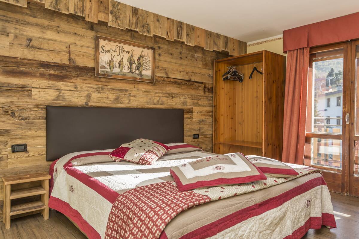 A bedroom with a wooden headboard, a bed with multiple quilts, a small bedside table, and a wardrobe.