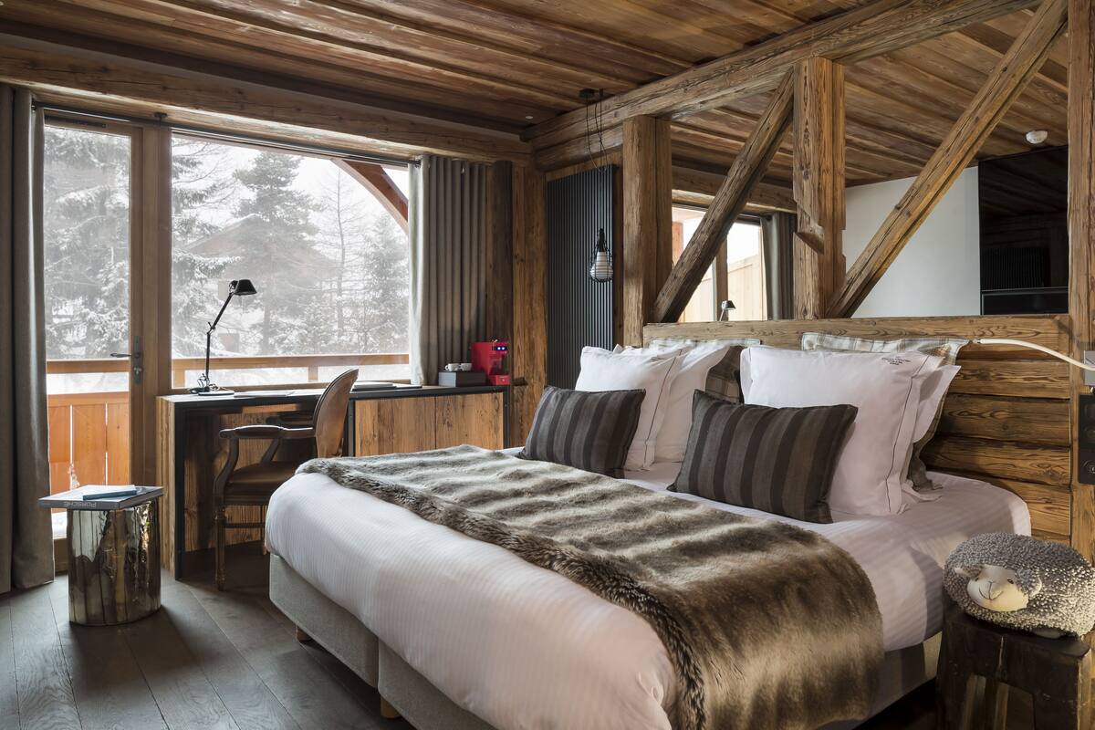 Un dormitorio acogedor con una cama grande, vigas de madera, un escritorio y vistas a árboles cubiertos de nieve.