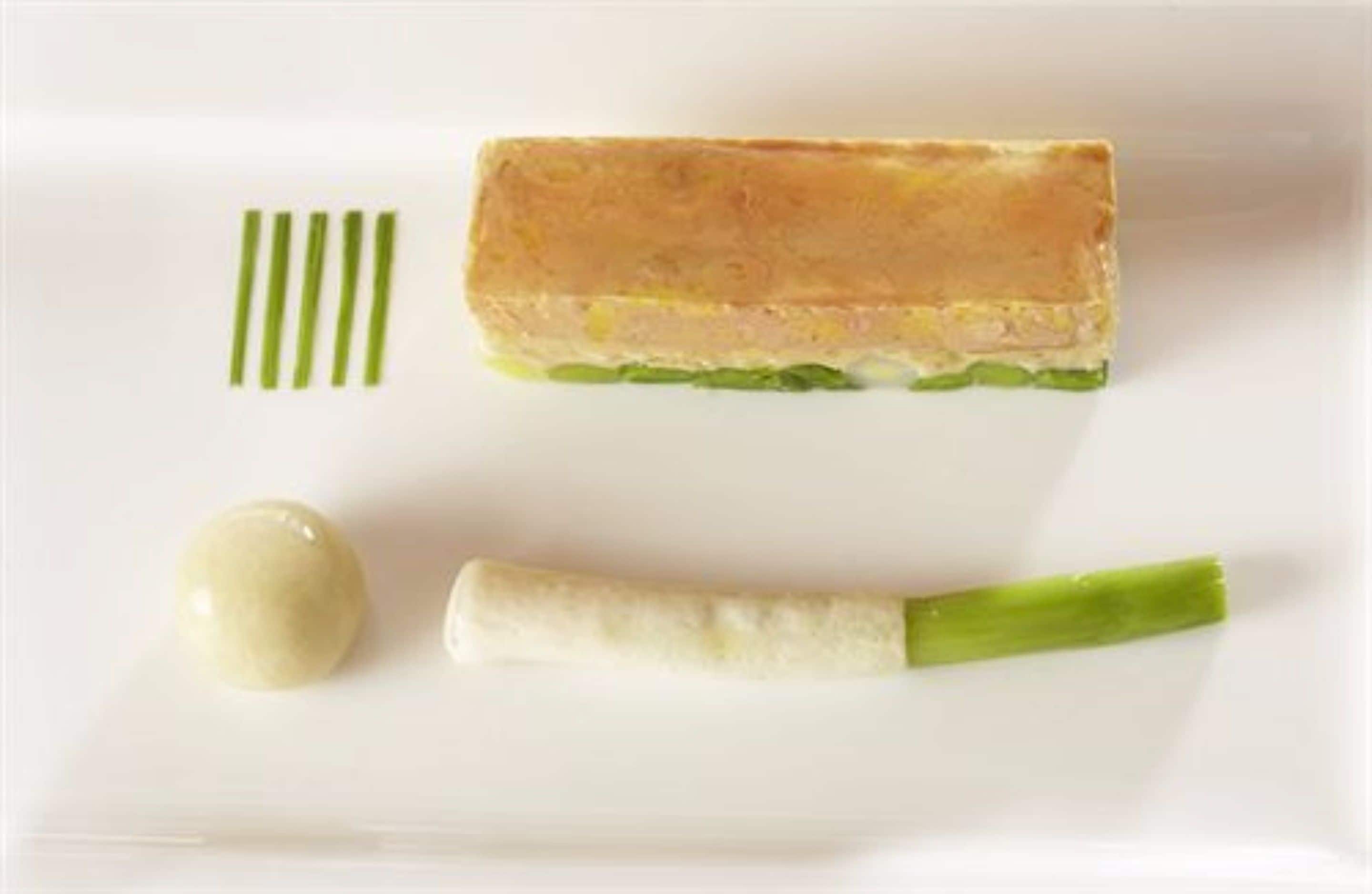 A rectangular dish with a golden-brown top layer, a green vegetable layer, and a white creamy substance, accompanied by a small white sphere and a green vegetable piece.