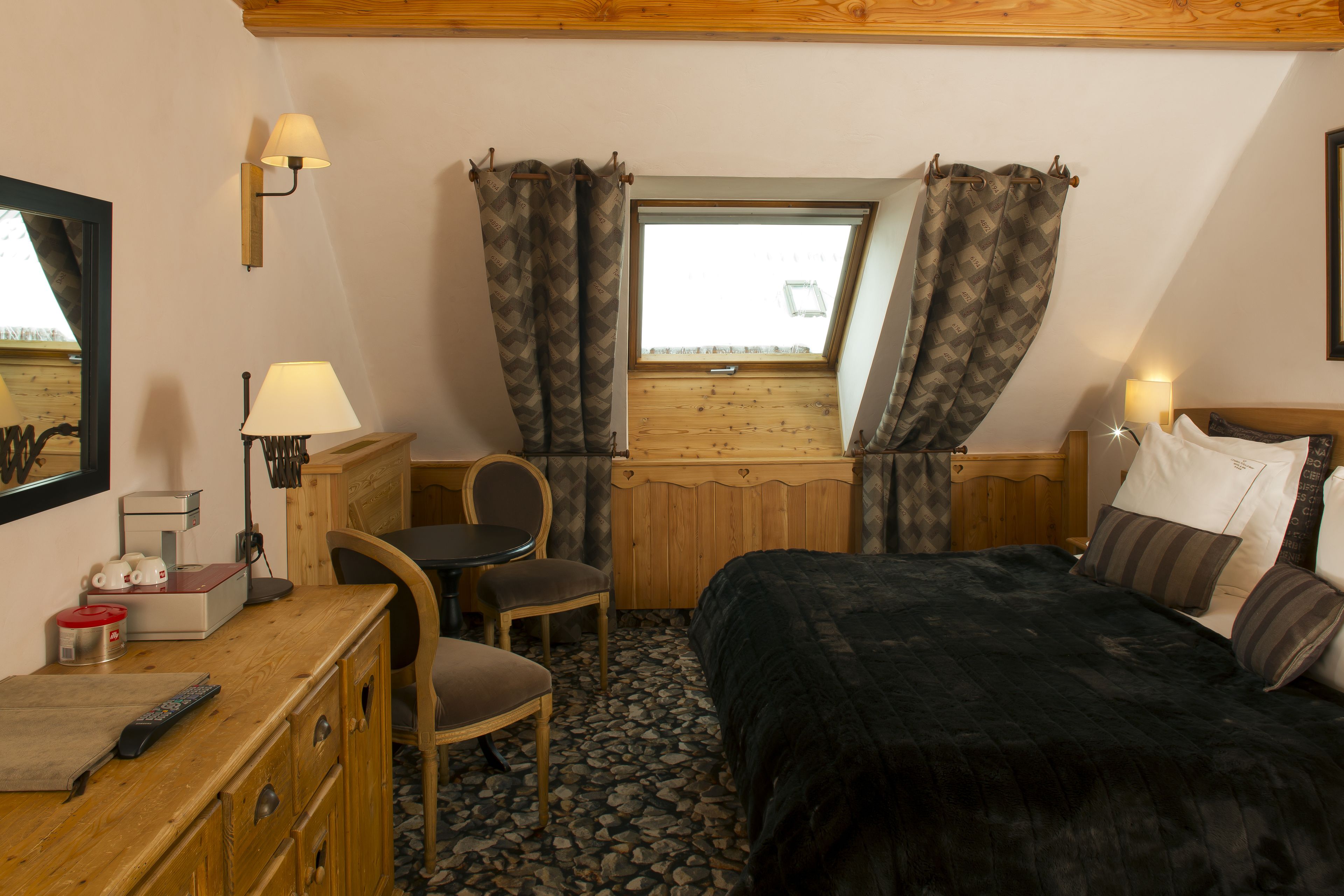 A compact hotel room with a bed, a desk with a chair, a small table with a lamp, and a window with curtains.