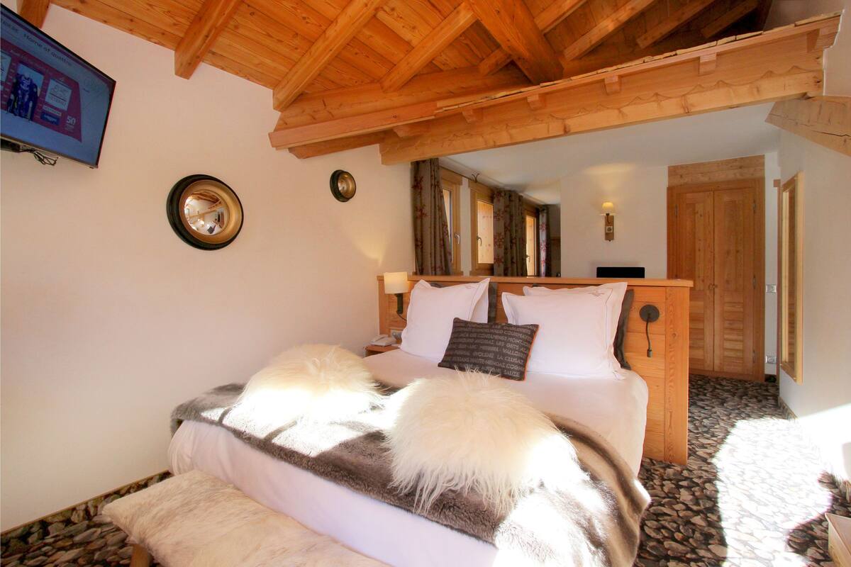 Un dormitorio con techo de madera, una cama con ropa de cama blanca y marrón, un espejo en la pared y un televisor a la izquierda.