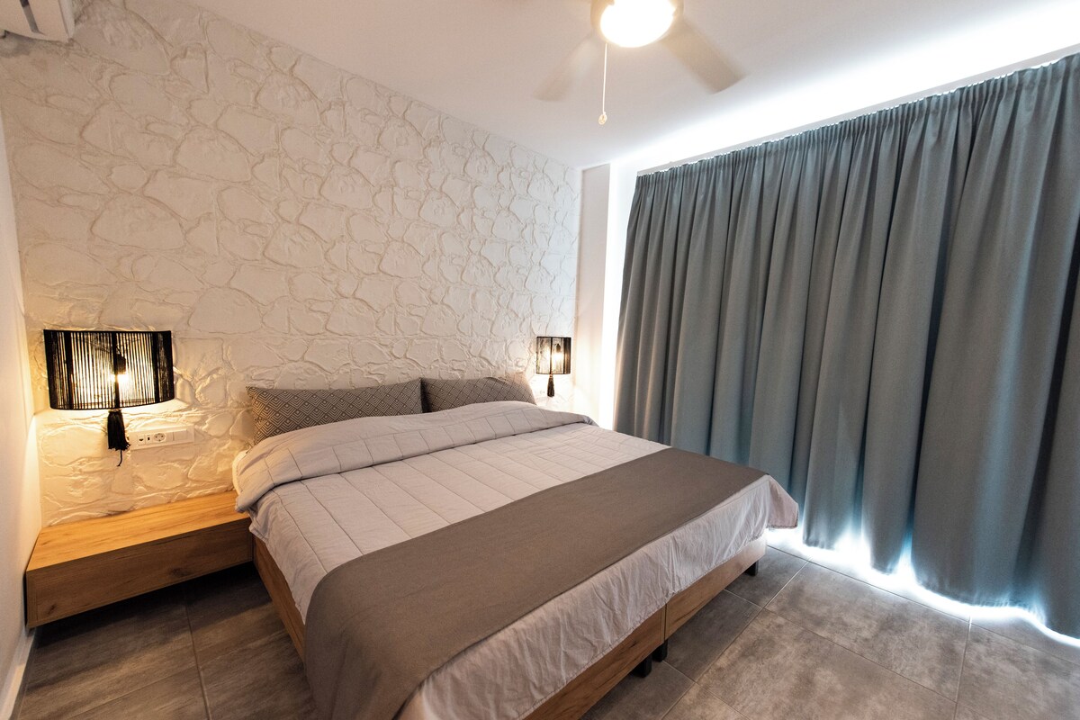 A bedroom with a stone wall, a bed with a headboard, a bedside table with a lamp, and a window with curtains.