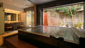 Un baño moderno con una bañera de hidromasaje con mampara de vidrio, taburetes de madera y vistas a un jardín a través de amplias ventanas.