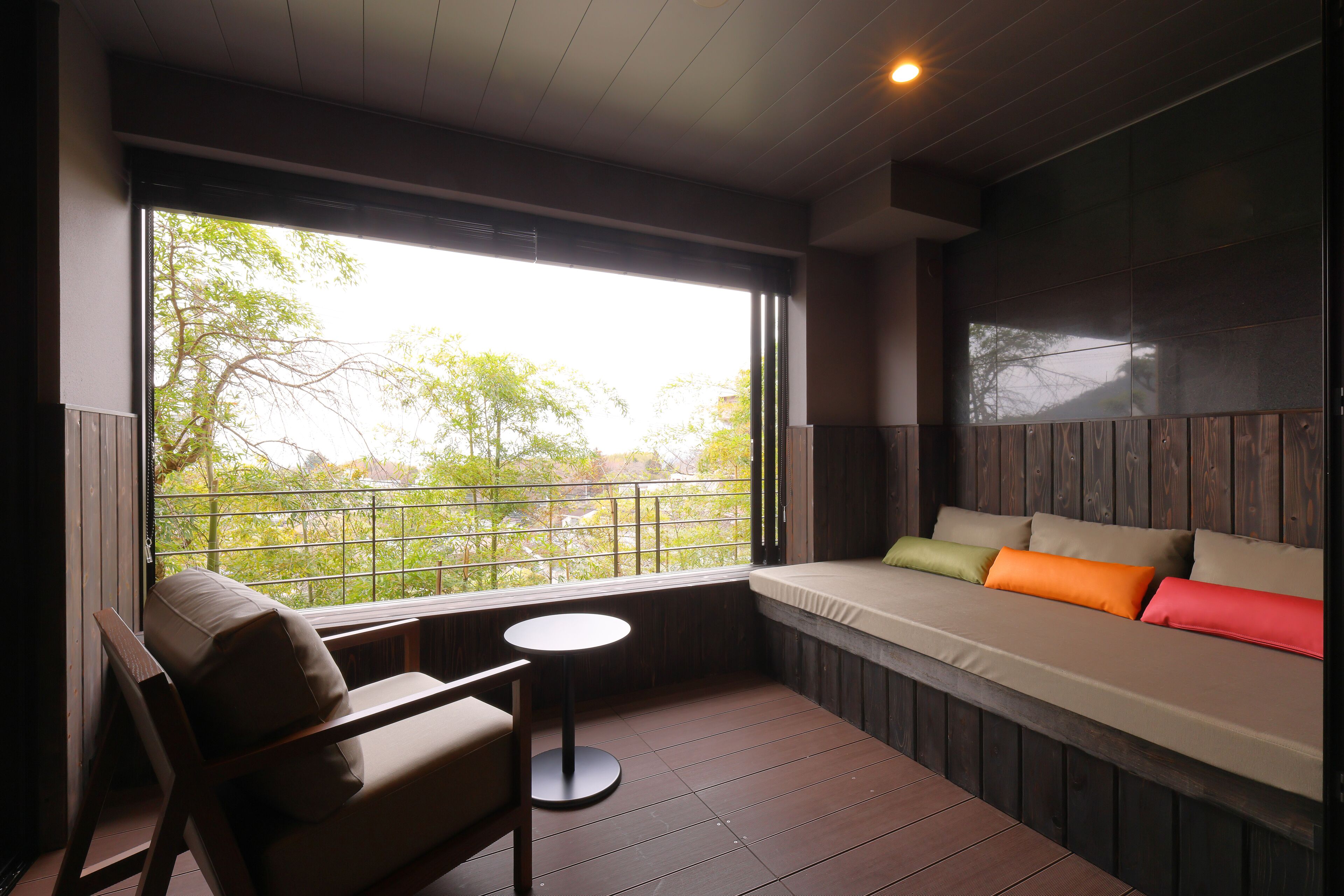A room with a wooden floor, a cushioned bench with pillows, a small round table, and a large window with a view of trees.