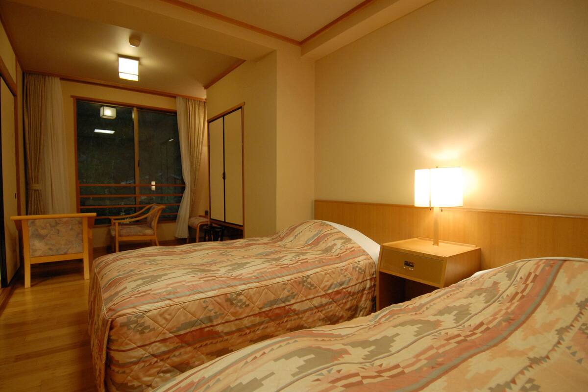 Habitación de hotel con dos camas, un escritorio, una silla y una mesita de noche con lámpara.