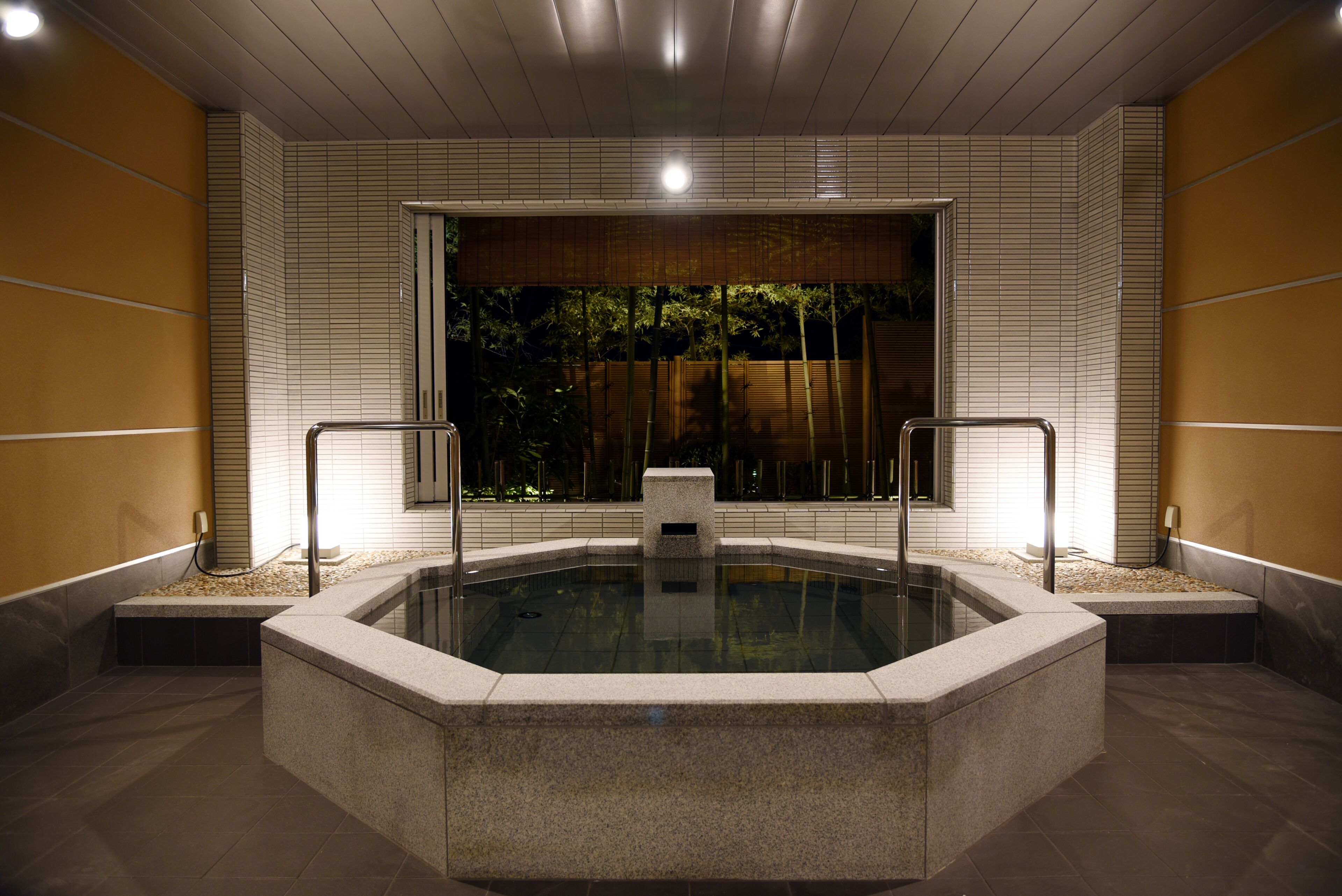A modern indoor swimming pool with a view of bamboo outside, surrounded by tiled walls and a wooden ceiling.