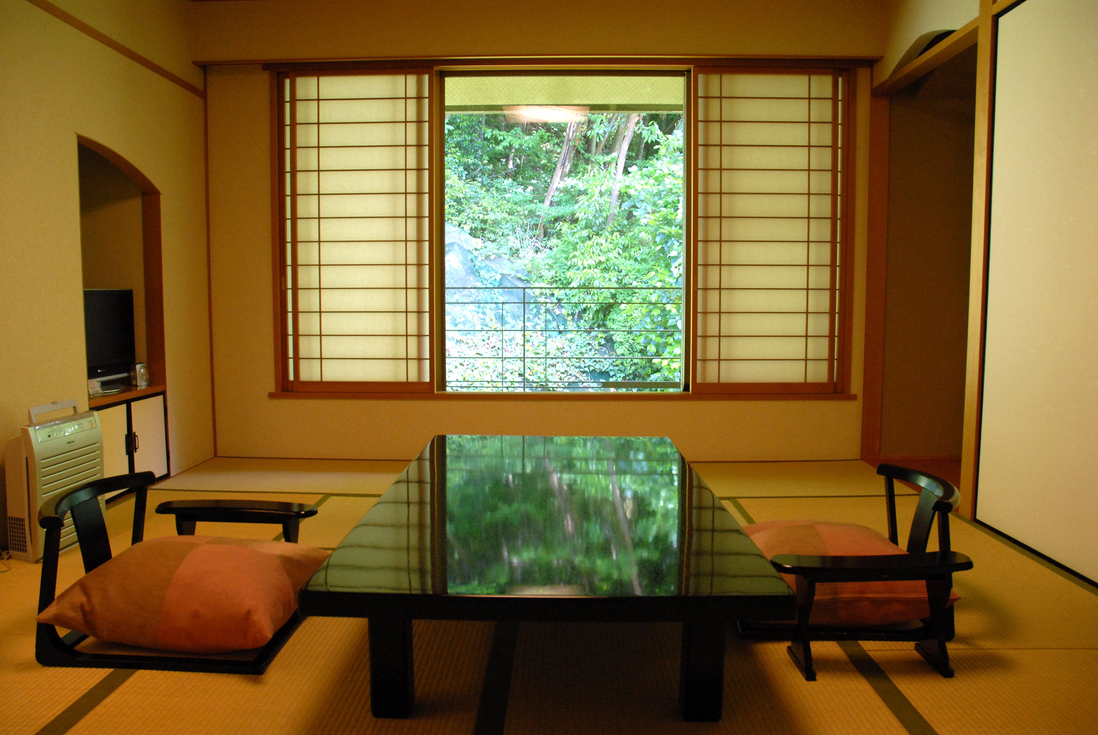 Una stanza con tatami, un tavolino, sedie, una televisione e vista su piante attraverso delle porte scorrevoli shoji.