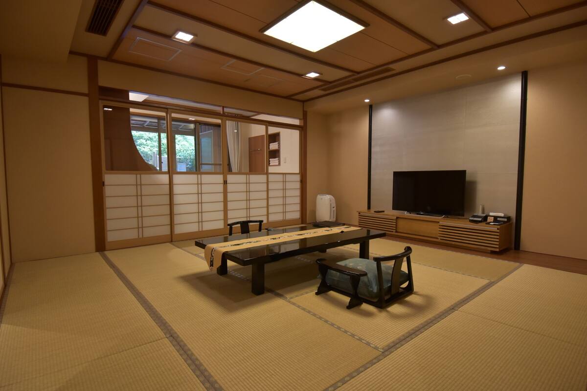Una habitación tradicional japonesa con suelo de tatami, una mesa baja, sillas, un televisor y puertas correderas.