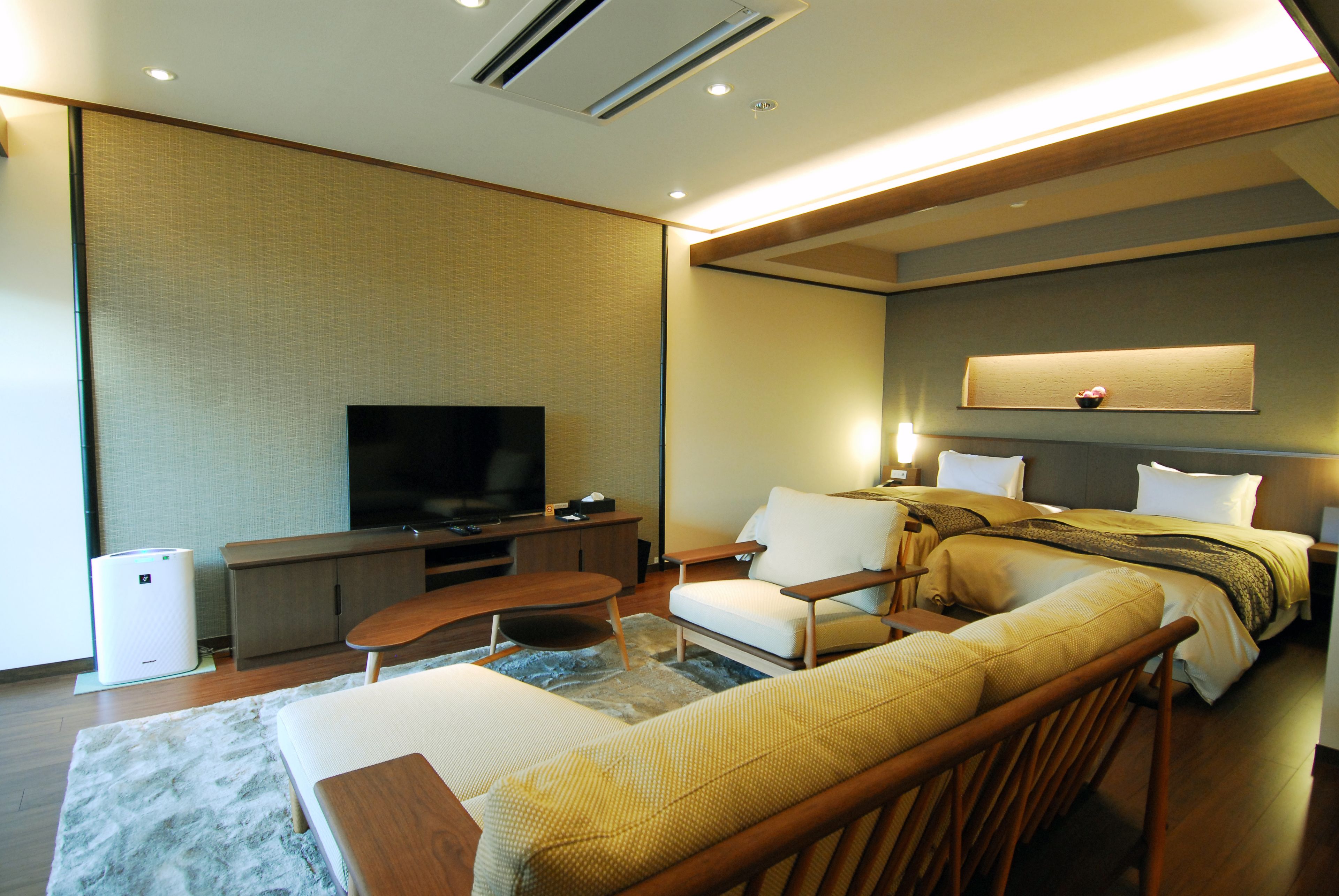 A modern hotel room with a flat-screen TV, a sofa, a chair, a bed, and a bedside table.