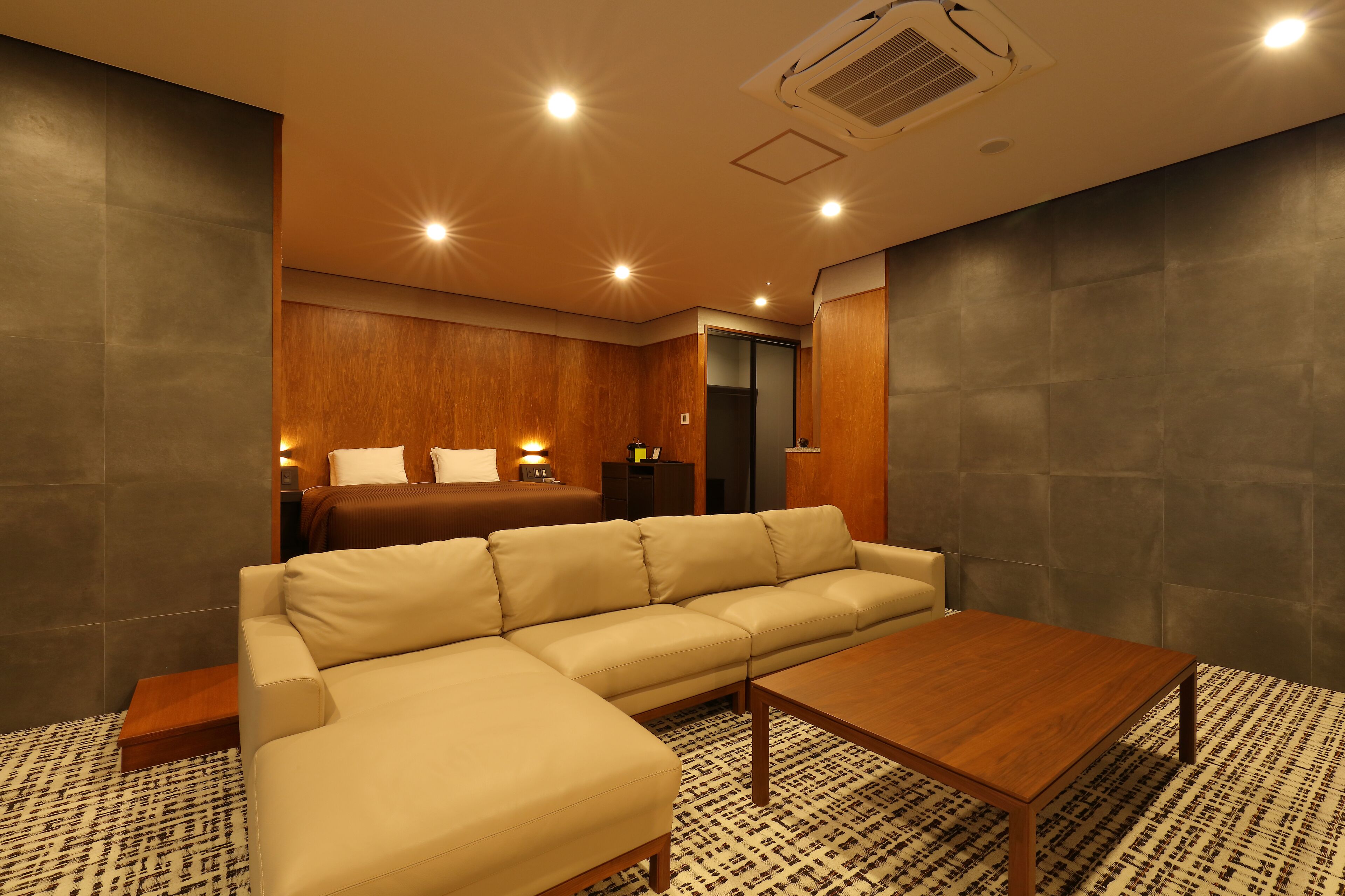A modern hotel room with a large beige sectional sofa, a wooden coffee table, and a patterned rug.