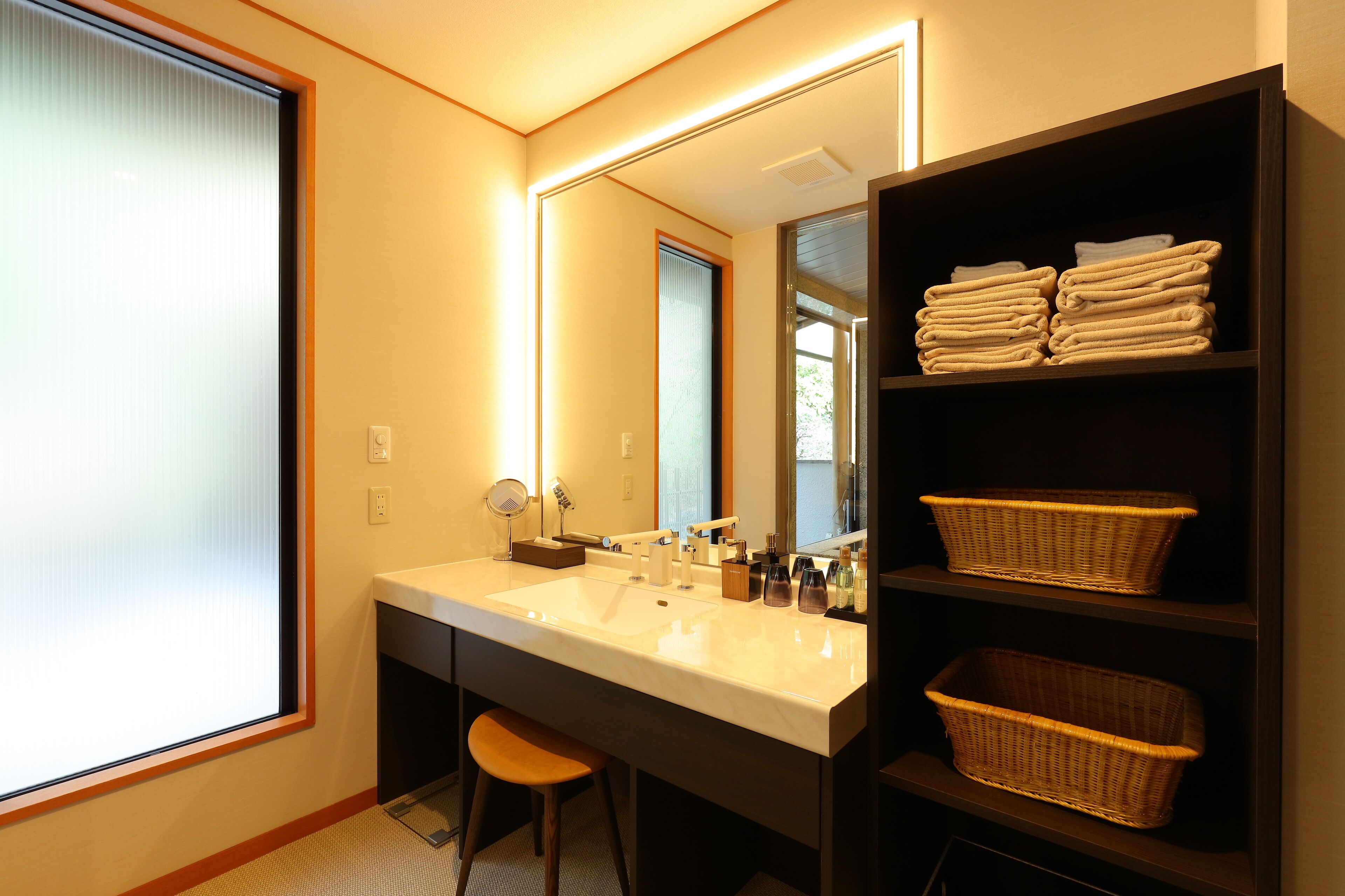 Une salle de bain moderne avec un grand miroir, deux lavabos, un tabouret et des paniers en osier placés sur des étagères.