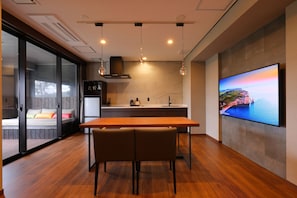 A modern kitchen with a wooden dining table, a TV mounted on the wall, and a sliding glass door leading to a balcony.