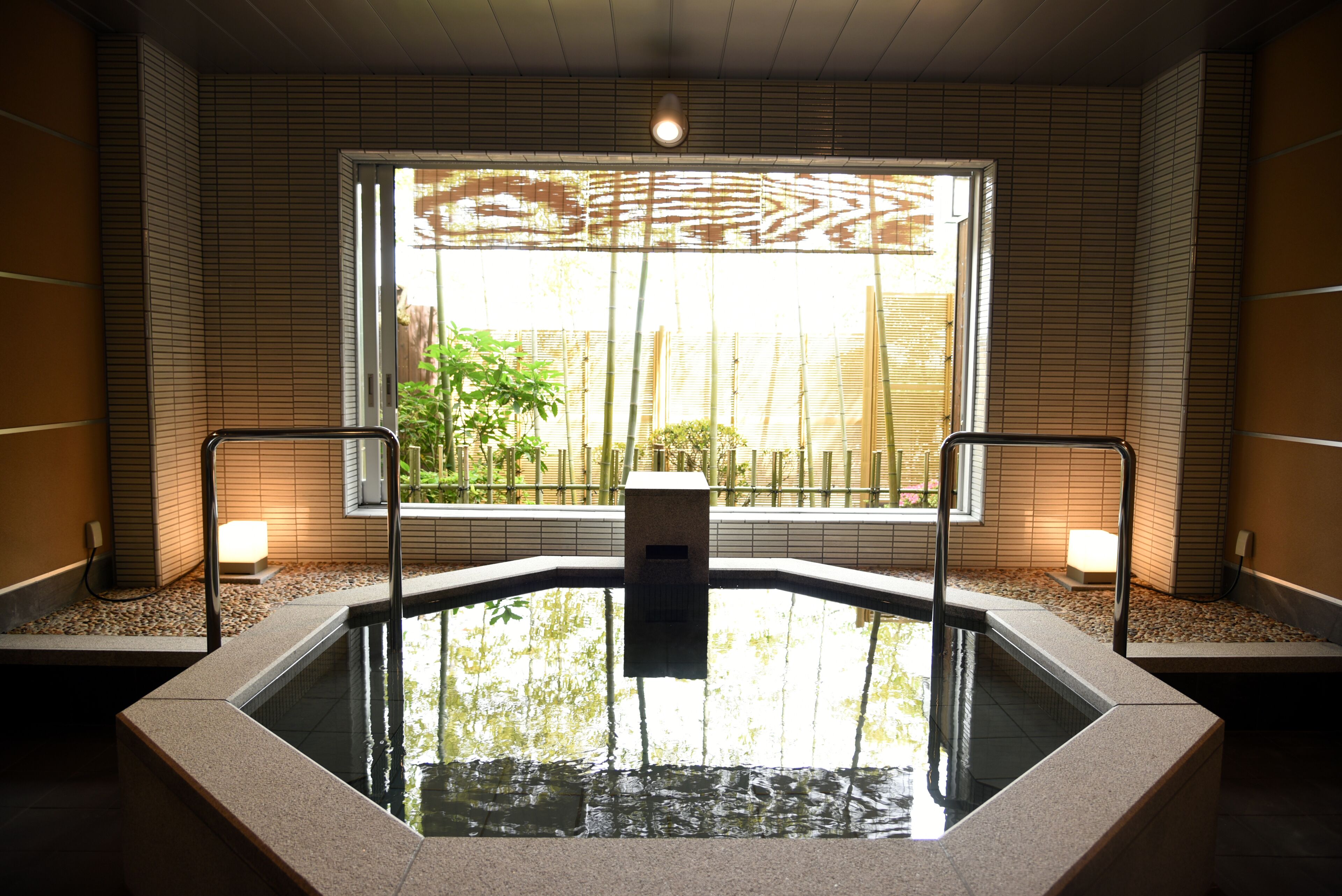 A hot tub with a view of bamboo outside, surrounded by tiled walls and a window.