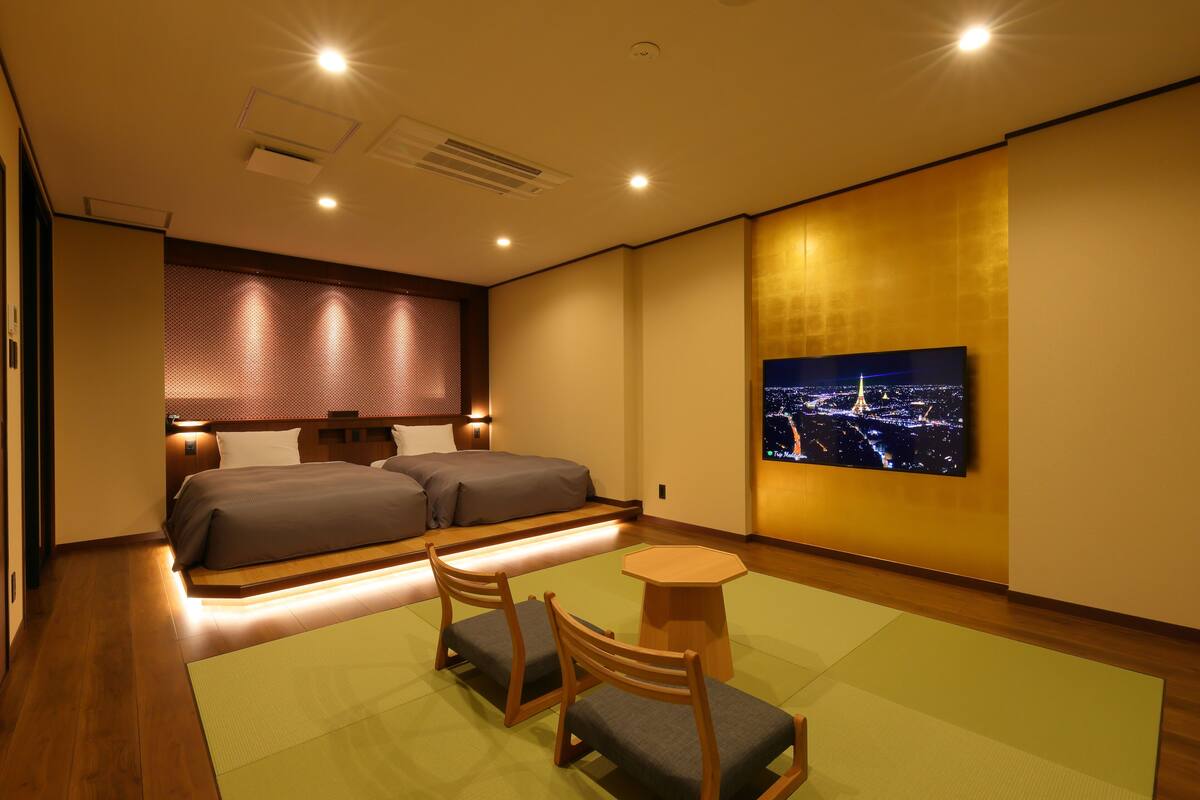 Habitación de hotel con una cama, dos mesitas de noche, una zona de estar pequeña con una mesa y sillas, y un televisor grande en la pared mostrando un paisaje urbano.