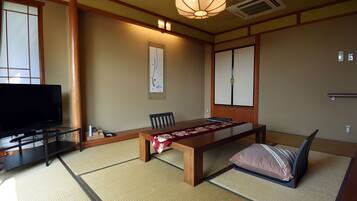 A traditional Japanese-style room with tatami flooring, a low table with a red and white patterned table runner, a television on a stand, and a sliding door.