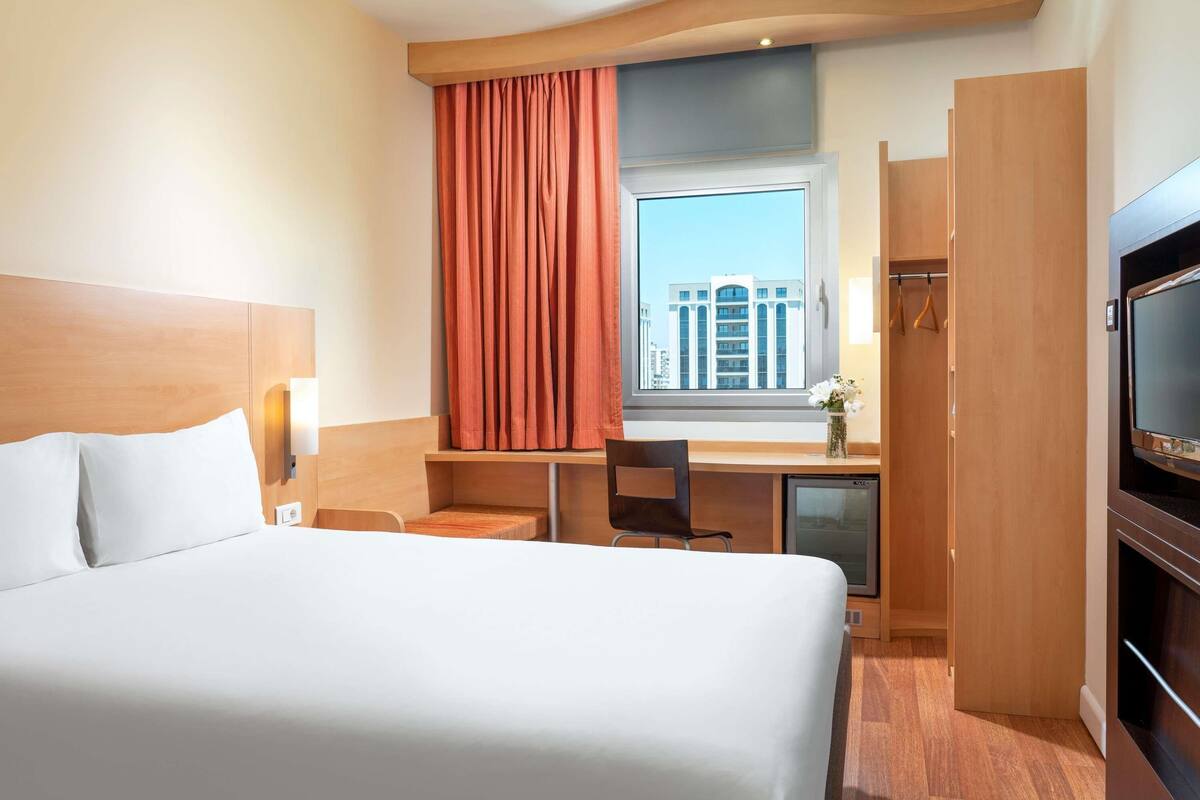 A hotel room with a large bed, a desk, a television, a window with curtains, and a view of a building.